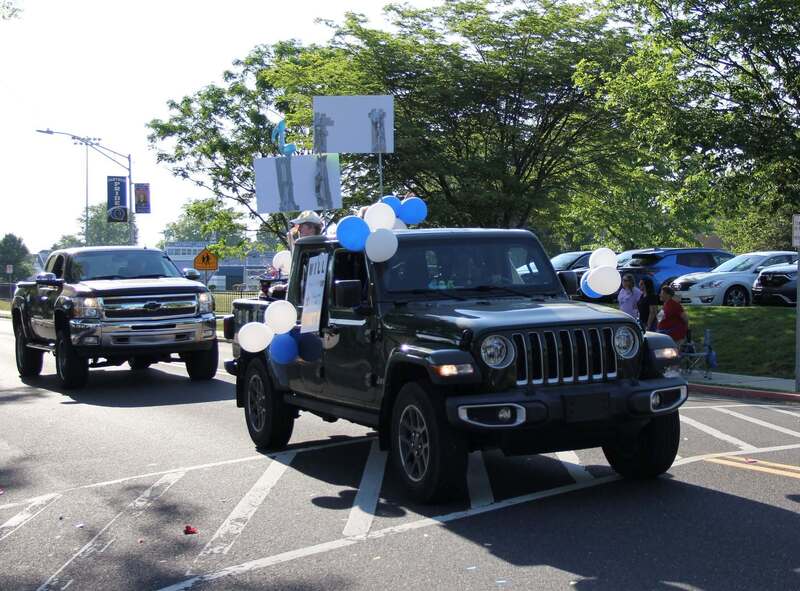 A decorated jeep with blue and white ballons drives slowly down the street