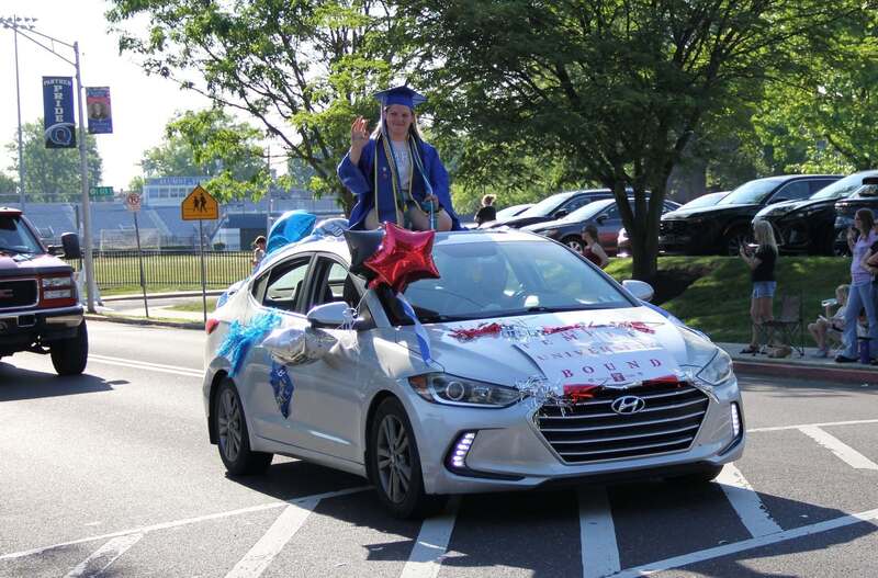 Students stands through cars sun roof in her cap and gown to wave to the crowd