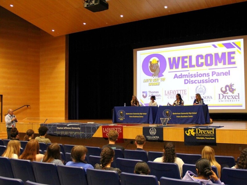 Four representatives sit up on a stage, seated behind a table. Behind them is a large projected image "Welcome Admissions Panel Discussion"
