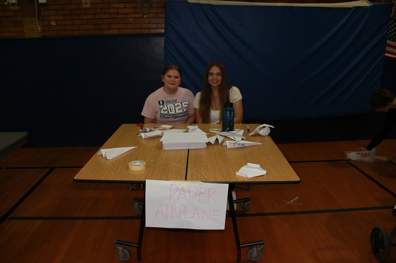 Two students sit behind a table at the Paper Airplane Station 