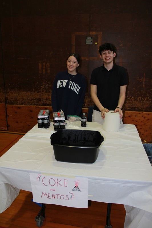 Coke and mentos station. Two students are standing behind a table with a white table cloth and black bucket to do their mentos and coke explosions in