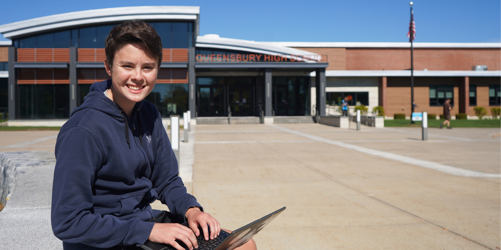 Ryan Greenstein sits in front of Queensbury High School with laptop.