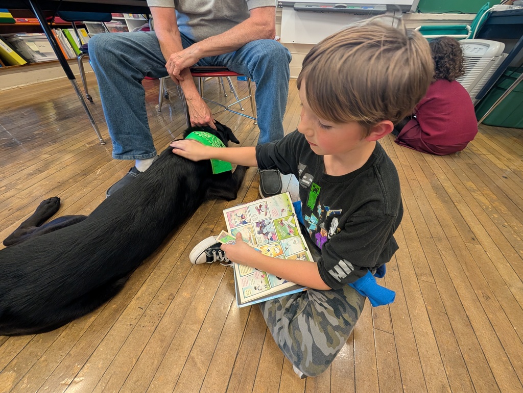 Pepper relaxes on the floor while a male student reads a book and pets Peppers shoulder.