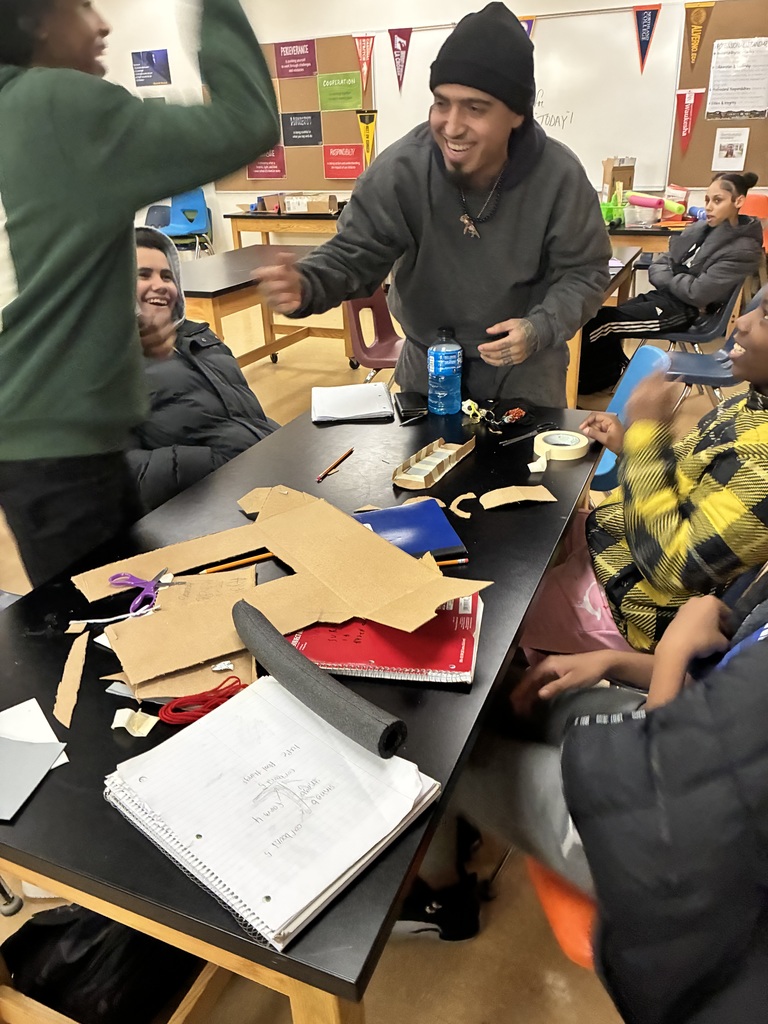 A smiling adult at a long black table, surrounded by middle schoolers. There's notebooks, scissors, masking tape, and scissors laying across the table.