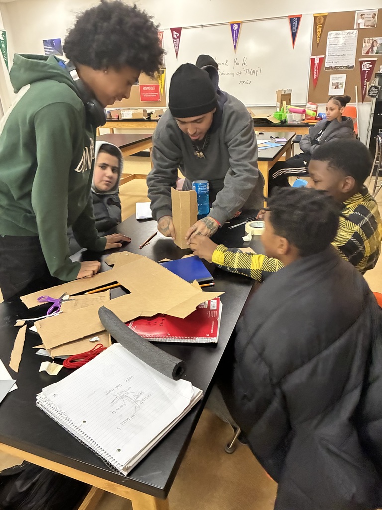 An adult at a long black table, surrounded by middle schoolers. There's notebooks, scissors, masking tape, and scissors laying across the table.