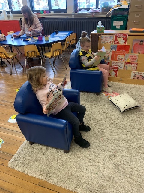 Two preschool girls sitting in comfortable chairs, reading books.