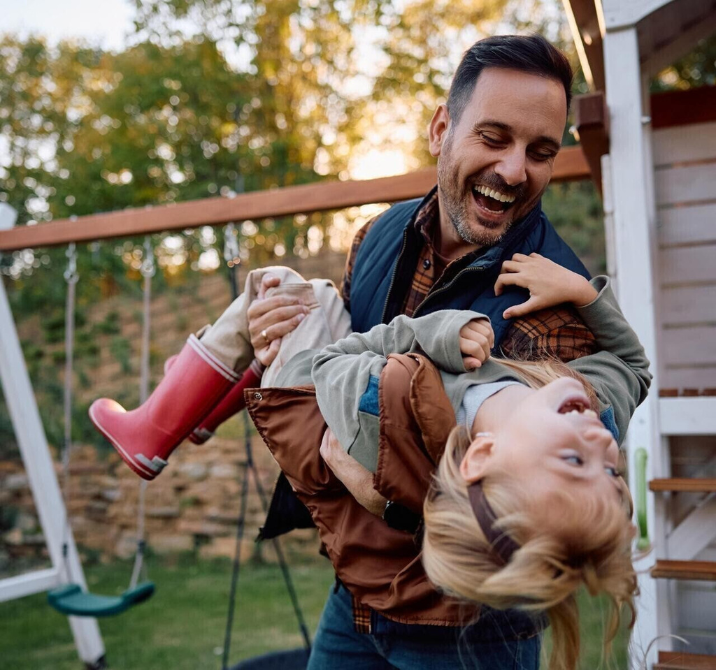 Photo of an adult male carrying a laughing young child while playing in a backyard.