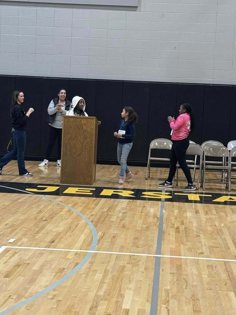The final two contestants are just finishing up the bee clapping for one another with the principal and a teacher standing behind them and the emcee at the podium.