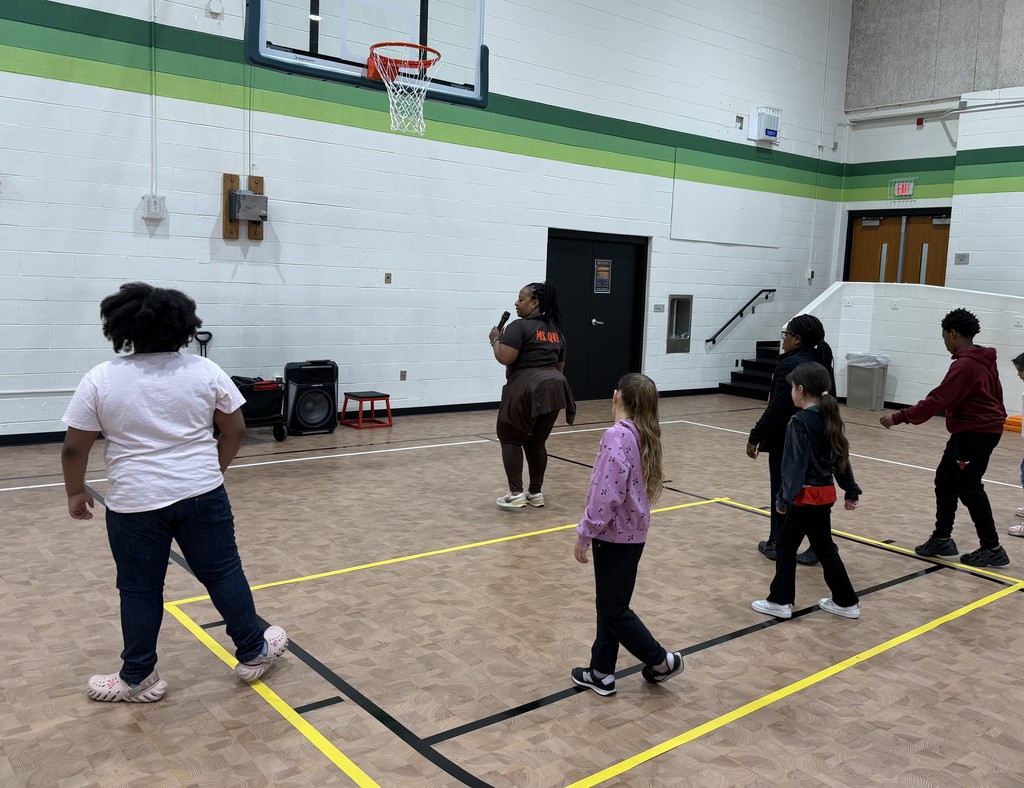 A group of people facing a white and green striped wall. A woman with a microphone is standing closest to the wall, guiding the students in a line dance.