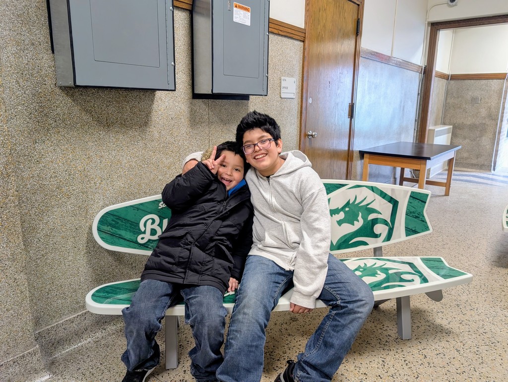 Two young boys sit on the bench. The boy on the left holds up a peace sign and an open mouthed smile. The boy on the right is smiling.