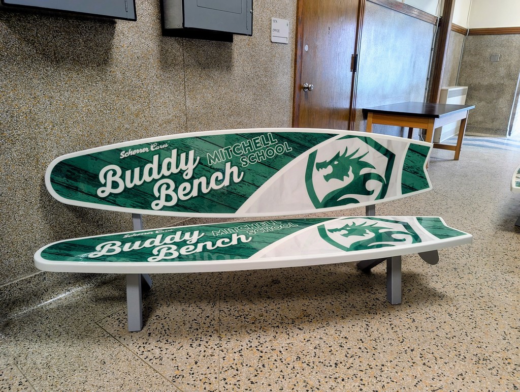 A bench resembling surfboards against a cement wall. It's green with white text that reads "Scherrer Cars Buddy Bench Mitchell School" on both the back rest and the seat. Beside the text is the Mitchell KnightDragon logo. It's in the shape of a shield with a dragon inside.