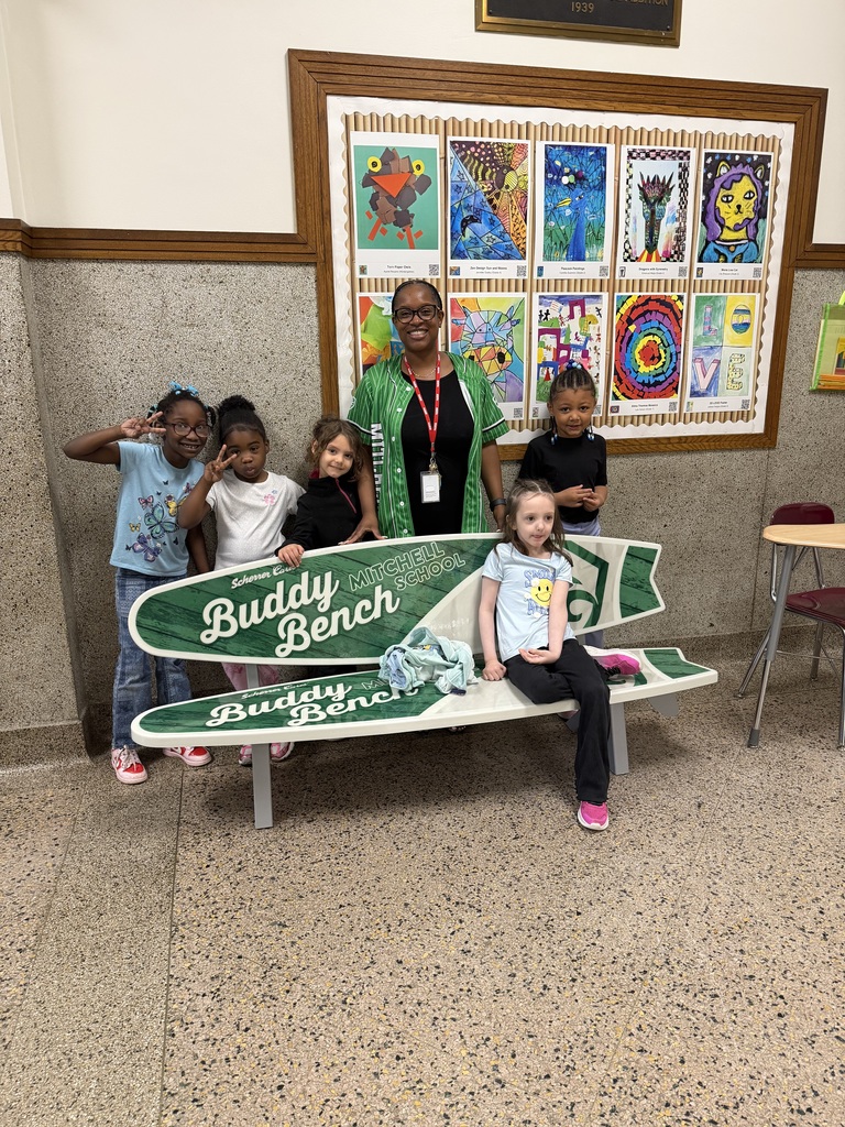 One elementary school girl sits on the bench with four classmates and a staff member standing behind it the buddy bench.