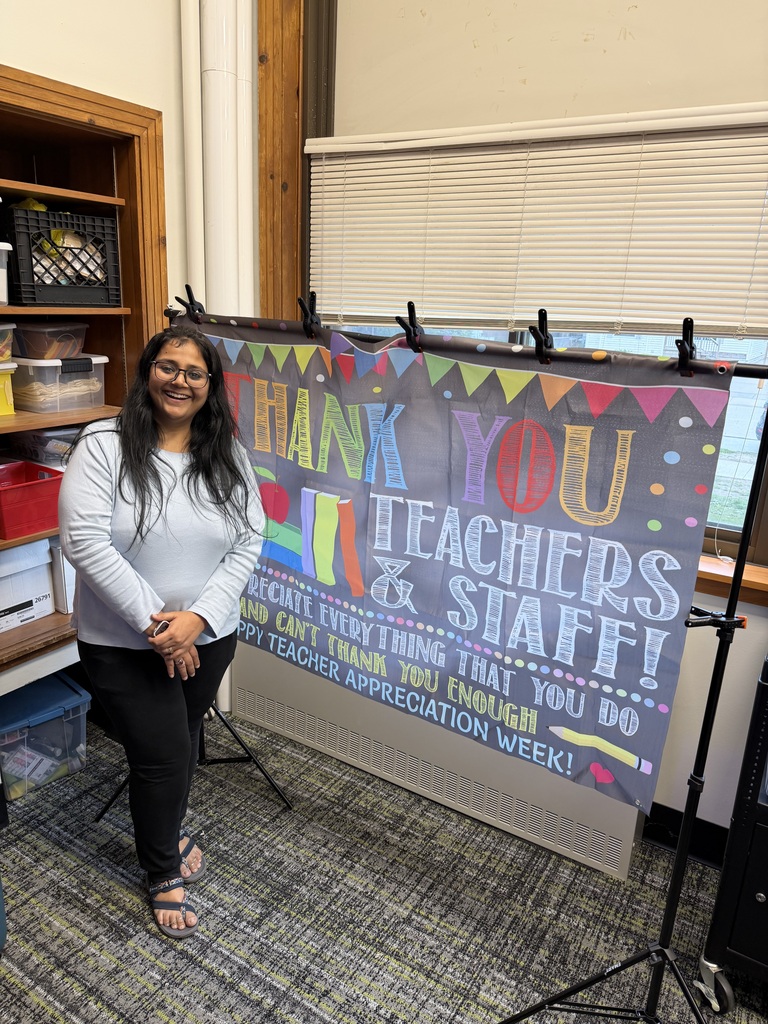 A teacher standing beside a sign that says "Thank you teachers & staff. We appreciate everything that you do and can't thank you enough. Happy teacher appreciation week!"