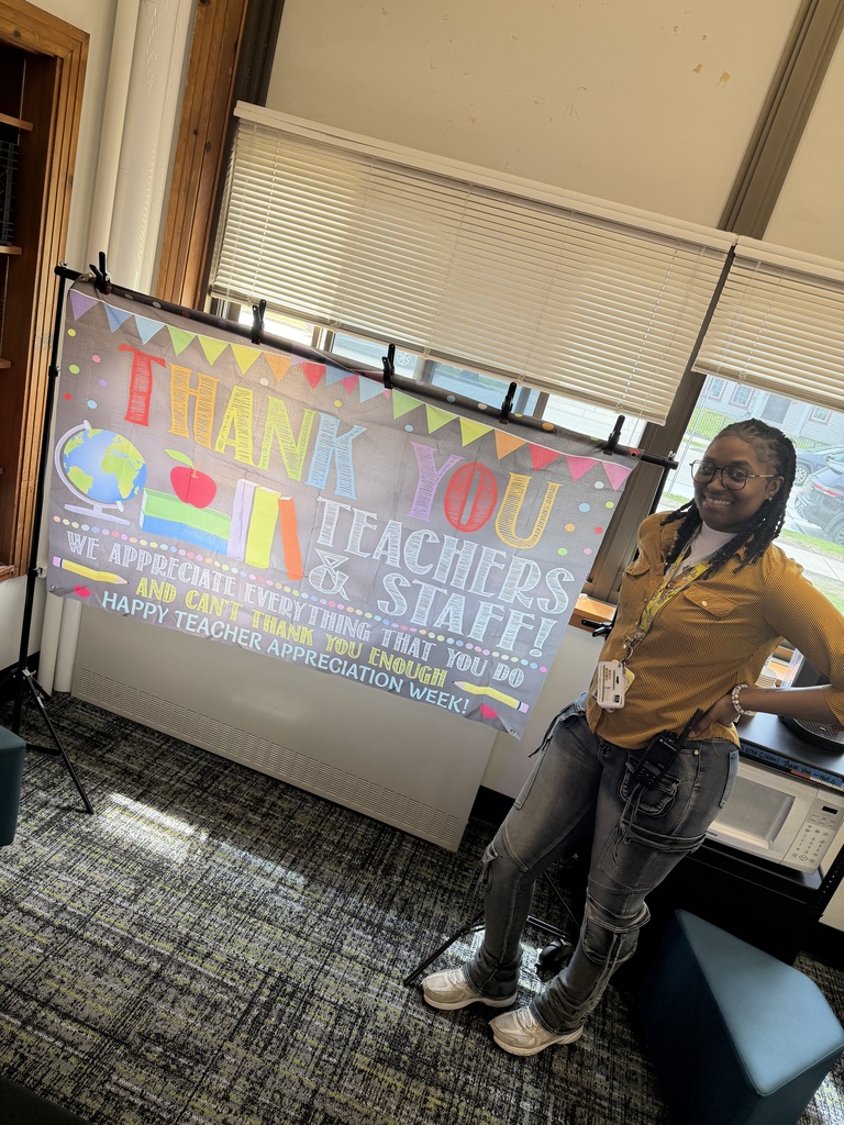 A teacher standing beside a sign that says "Thank you teachers & staff. We appreciate everything that you do and can't thank you enough. Happy teacher appreciation week!"