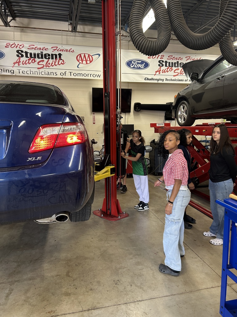 Three Mitchell students watch a car lift lower a car.