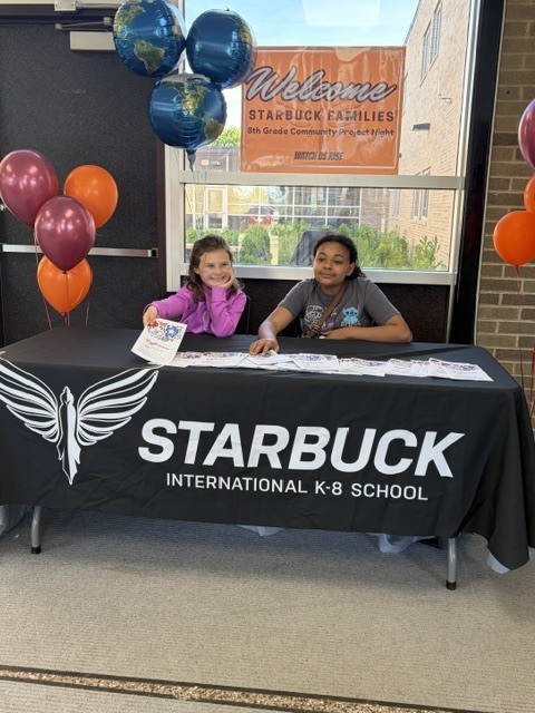 Two students sitting at a table with a Starbuck International K-8 School logo, handing out programs.s