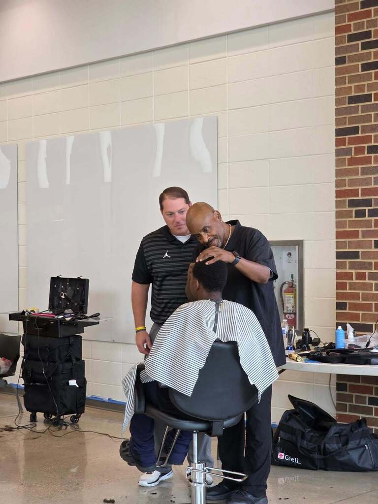 Directing principal looks on as Dr. G cuts a student's hair.