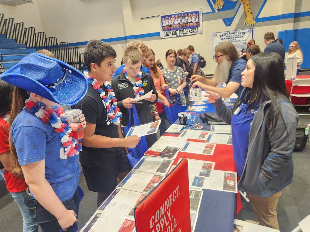 A group of boys dressed in red, white, and blue talk to a person at a booth for college and career fair 