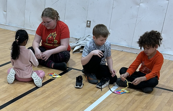 2 SGA Members and two younger students sitting on gym floor tying shoelaces.
