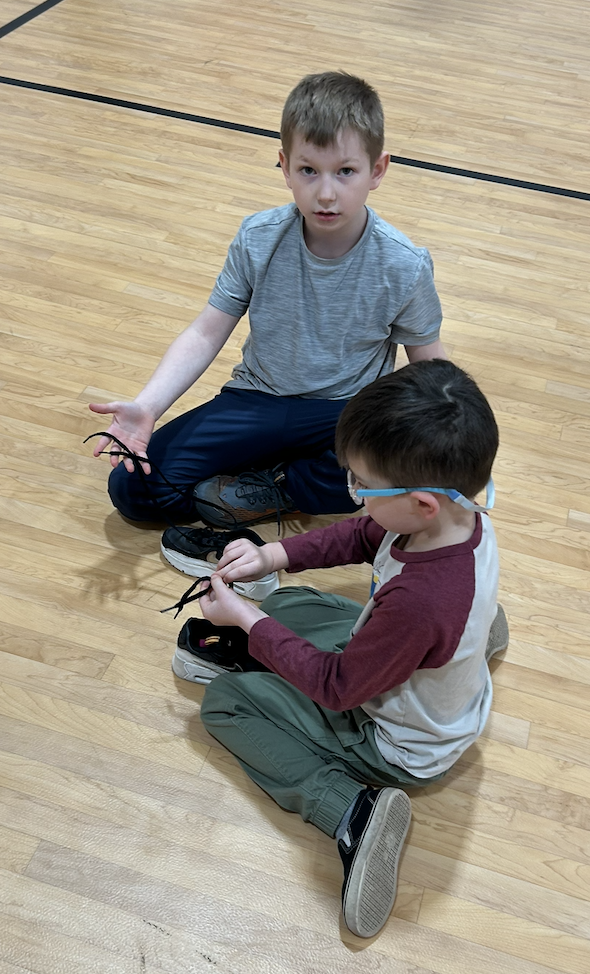 SGA Member and kindergarten student sitting on gym floor tying shoelaces.