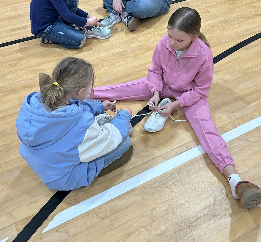 SGA Member and first grade student sitting on gym floor tying shoelaces.