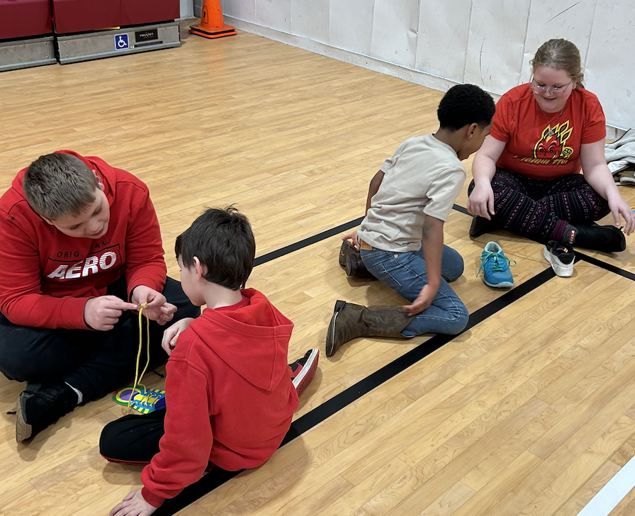 2 SGA Members and two younger students sitting on gym floor tying shoelaces.