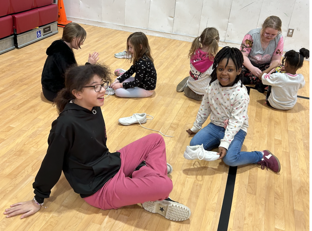 3  SGA Members and four younger students sitting on gym floor tying shoelaces.