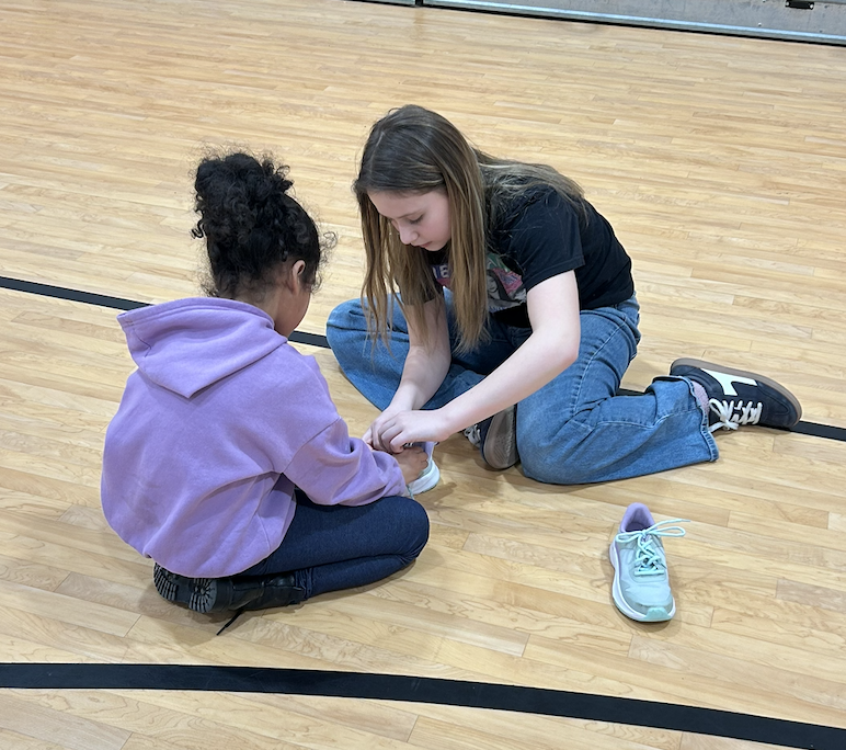 SGA Member and kindergarten student sitting on gym floor tying shoelaces.