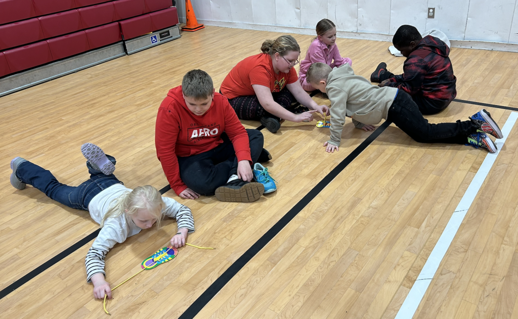3 SGA Members and three younger students sitting on gym floor tying shoelaces.