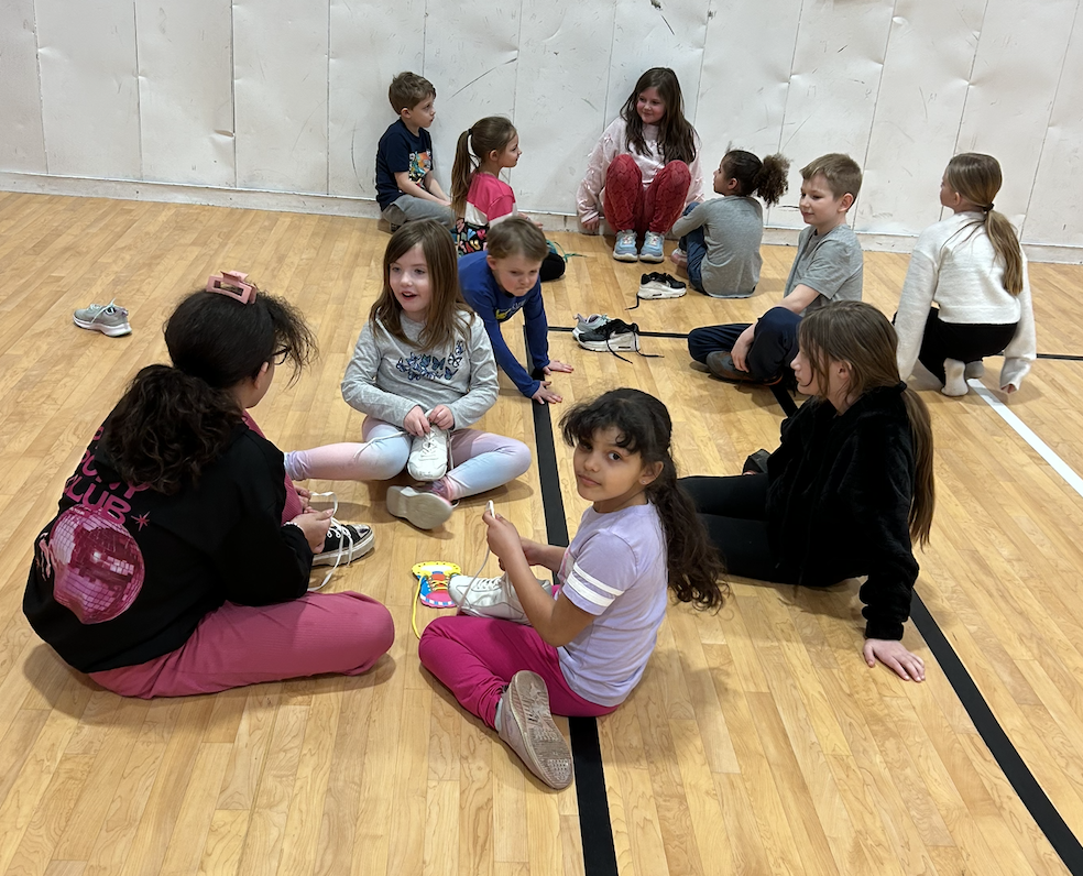 5 SGA Members and 6 kindergarten students sitting on gym floor tying shoelaces.