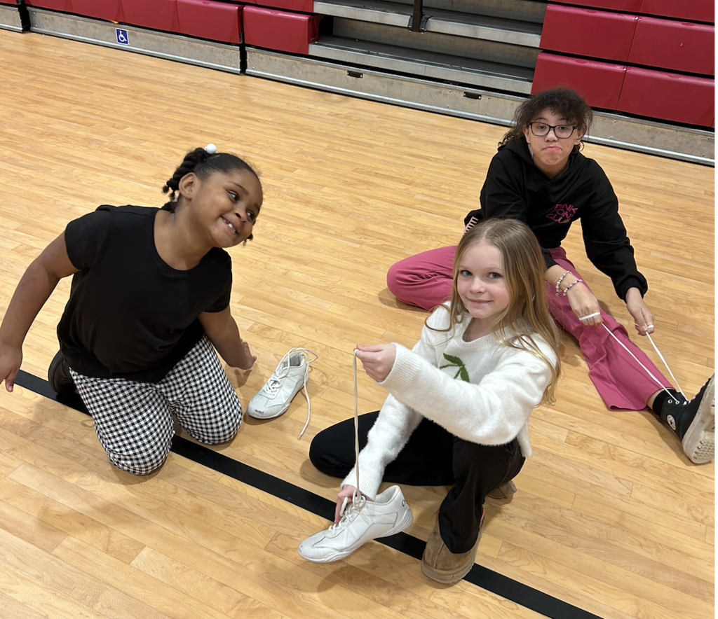 2 SGA Members and one younger student sitting on gym floor tying shoelaces.