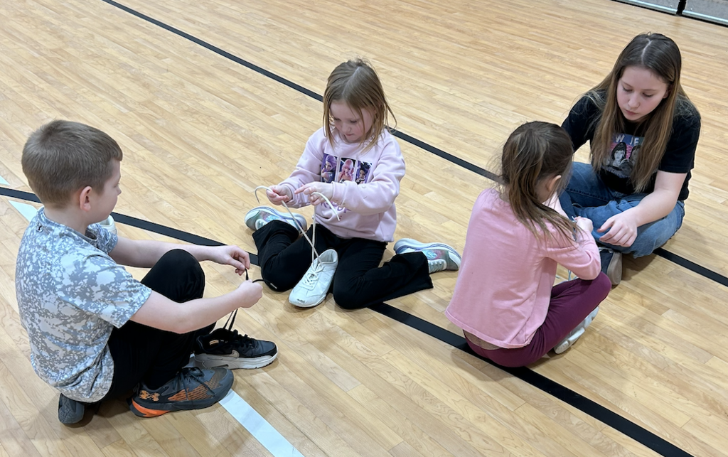 2 SGA Members and two younger students sitting on gym floor tying shoelaces.