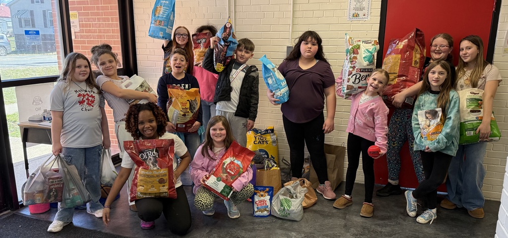 SGA Pet Supply Drive- Elementary students smiling and holding donated food and pet supplies inside a school hallway.