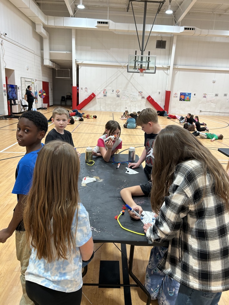 Several children stand around a black folding table in a gymnasium, working on a craft activity with markers, pipe cleaners, and small decorated paper pieces spread across the table. Some children are drawing or assembling materials, while others look toward the camera. In the background, additional children are seated or lying on the gym floor, and a basketball hoop and posters are visible on the far wall.
