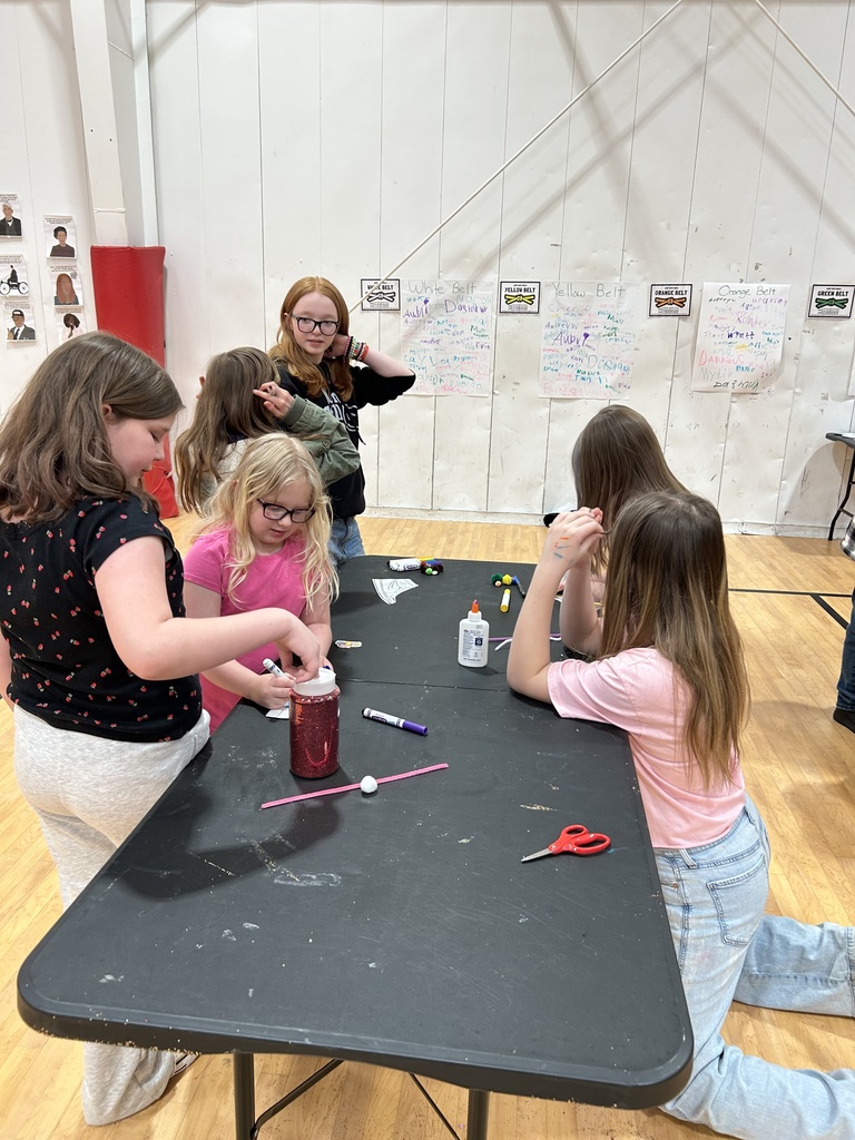 Several children are gathered around a black rectangular craft table in a gymnasium. Some children are seated while others stand as they work with various craft supplies, including glue bottles, markers, and a jar filled with red material. One child is reaching into the jar, while others watch or work on their own projects. In the background, additional children stand near the wall, which is covered with posters and handwritten charts. The gym floor and white walls are visible throughout the scene.