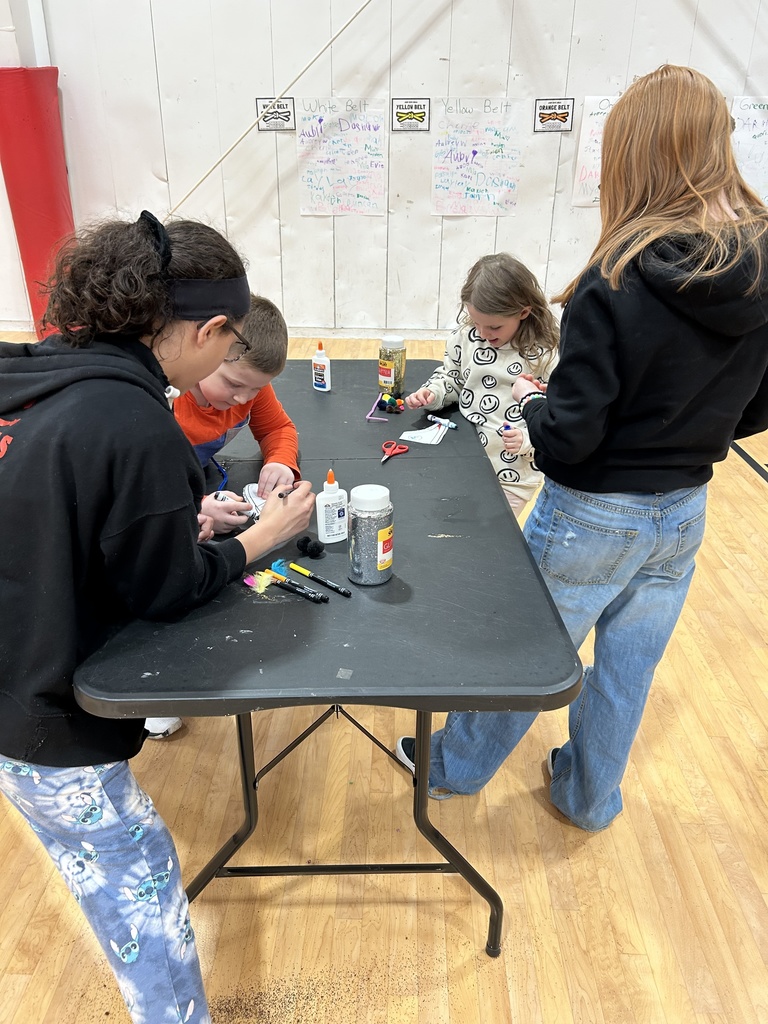 Four children are gathered around a black folding table in a gymnasium, working on a craft activity. The table is covered with glue bottles, scissors, markers, and small craft materials. Two children stand on the far side of the table while two lean over from the near side, all focused on their projects. Posters and signs are visible on the wall in the background.