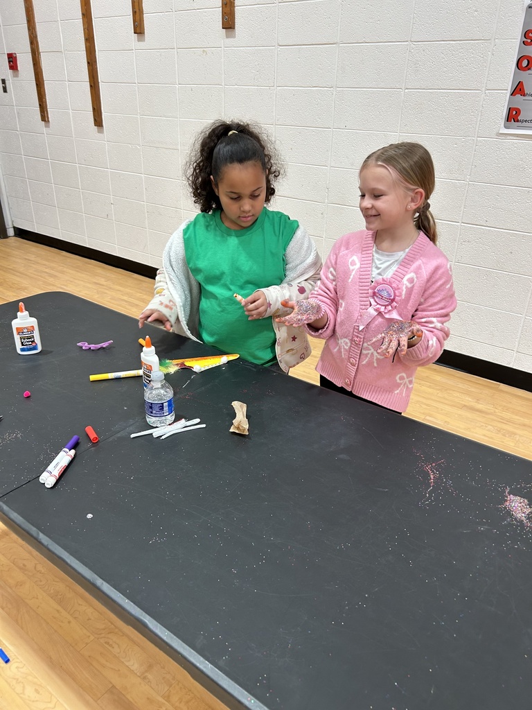 Two children stand at a black craft table in a gymnasium, working with glue and various craft supplies. The child on the left is wearing a green shirt with a light-colored sweater, and the child on the right is wearing a pink cardigan while holding craft materials. The table has scattered markers, glue bottles, and bits of glitter. A white brick wall and the gym floor are visible in the background.
