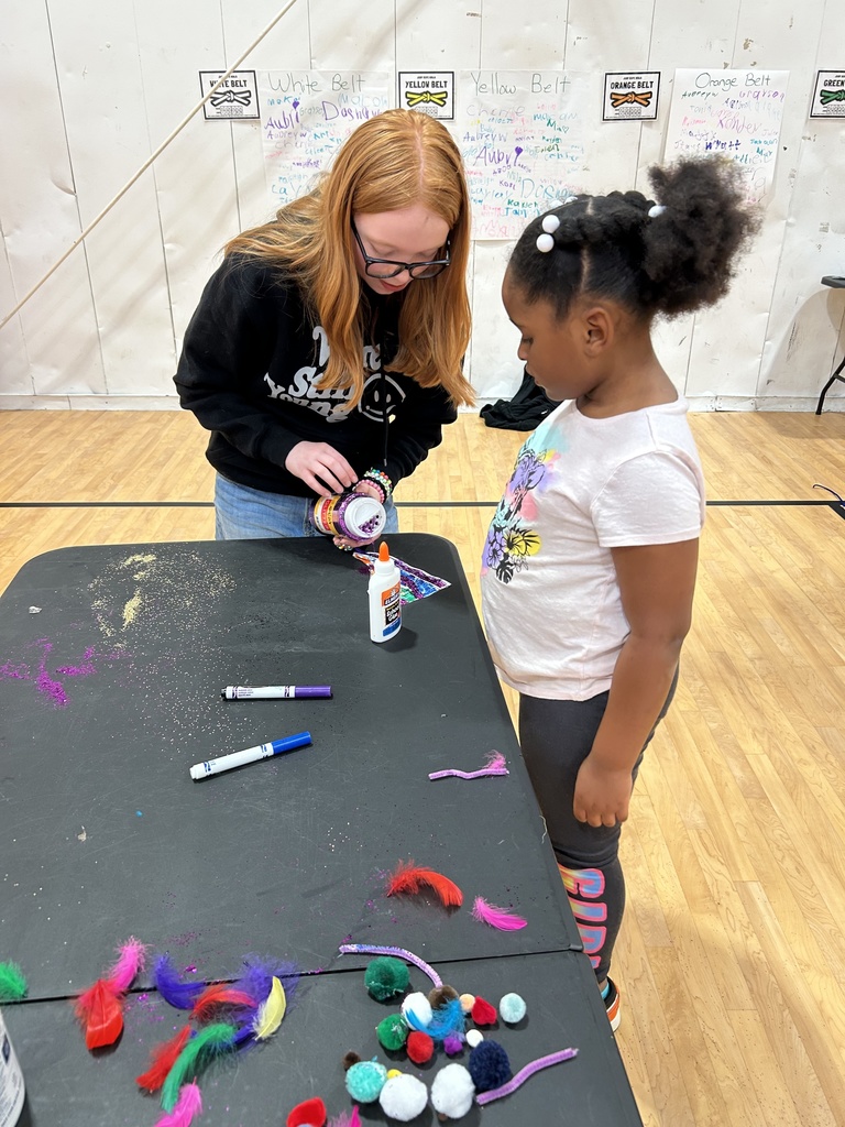 An older child stands at a black craft table in a gymnasium, assisting a younger child with a craft project. The older child is holding a circular craft item, while the younger child watches. The table is scattered with craft materials, including glue bottles, markers, colorful feathers, and pom‑poms. The gym floor, white walls, and posters in the background are visible.