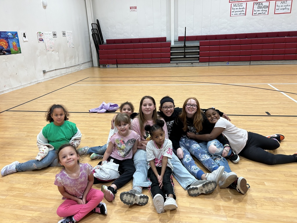 A group of children sit together on the floor of a gymnasium, arranged in two rows. The children in the front row sit cross‑legged, while those in the back row sit with legs extended. Behind them are red bleachers, a wall with posters, and the gym’s hardwood floor. The group is centered in the middle of the gym, posing closely together.