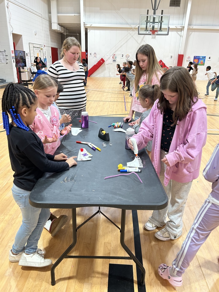 A group of children stand around a black rectangular table in a gymnasium, working on a craft activity. Various supplies such as glue bottles, markers, pipe cleaners, and small craft items are spread across the table. An adult stands behind the group, observing their work. More children and activity stations can be seen throughout the gym in the background, along with a basketball hoop and posters on the walls.