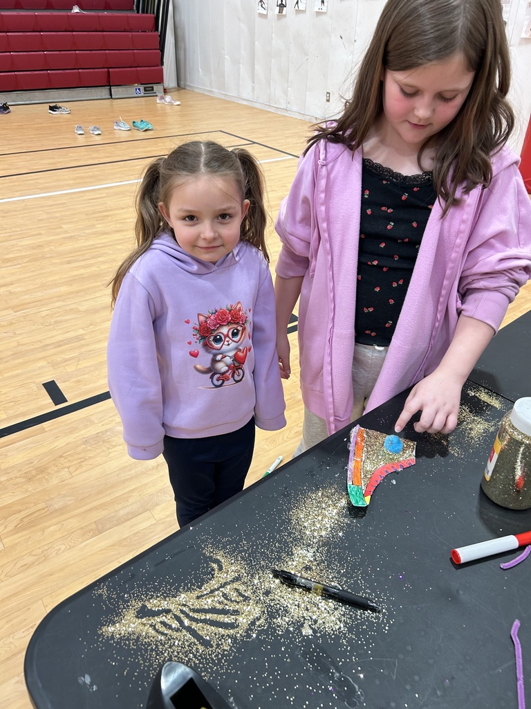 Two children stand beside a black craft table in a gymnasium. One child is wearing a light purple hoodie with a cartoon graphic on the front, and the other is wearing a pink jacket over a black shirt. On the table in front of them is a decorated paper craft covered in glitter, along with scattered glitter, markers, and a container of glitter. Red bleachers and shoes on the gym floor are visible in the background.
