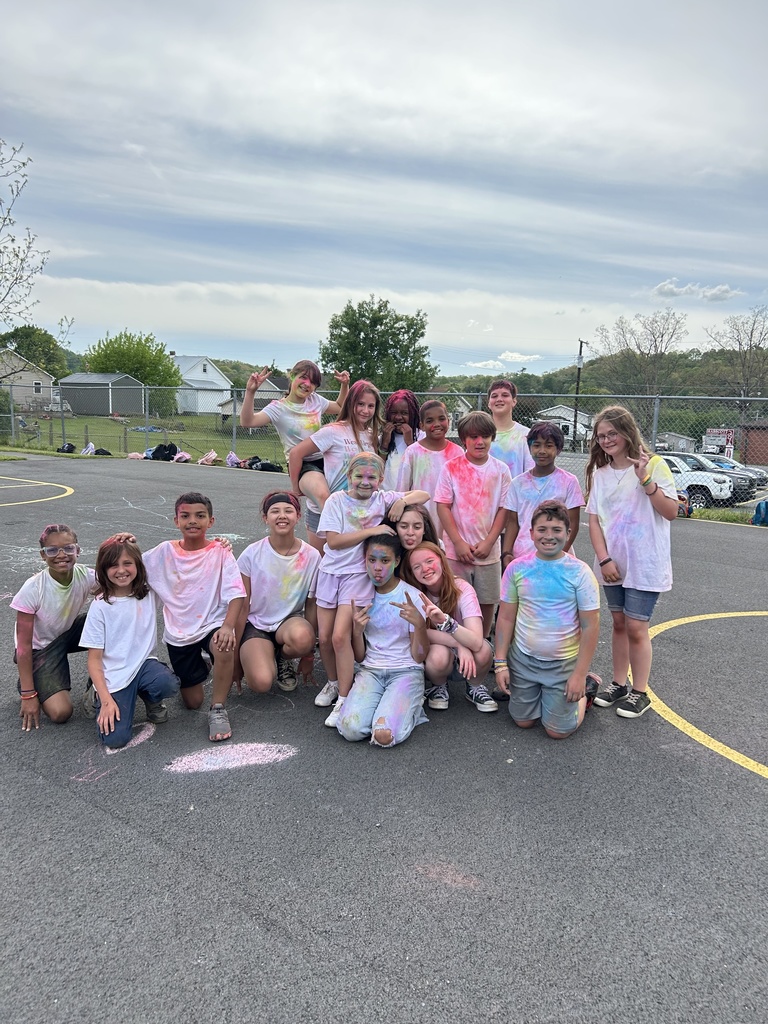 A group of elementary students stand in a line on an outdoor playground blacktop, smiling and posing with colorful powder on their white shirts. Chalk drawings are visible on the pavement in front of them, with trees, playground equipment, and a blue sky in the background.