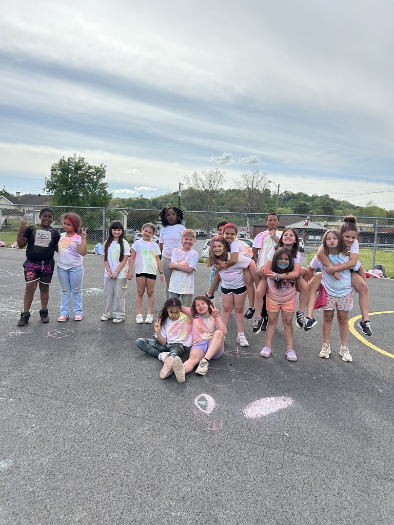 A group of elementary students stand in a line on an outdoor playground blacktop, smiling and posing with colorful powder on their white shirts. Chalk drawings are visible on the pavement in front of them, with trees, playground equipment, and a blue sky in the background.