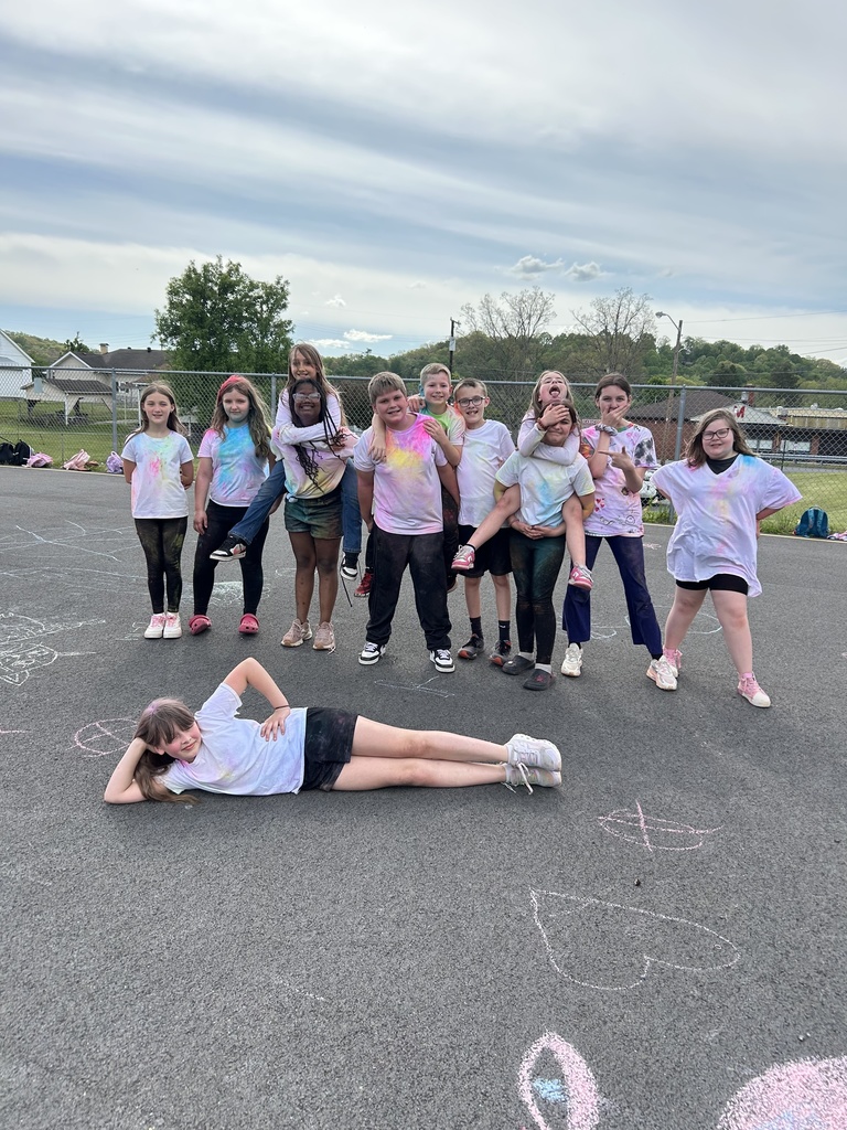A group of elementary students stand in a line on an outdoor playground blacktop, smiling and posing with colorful powder on their white shirts. Chalk drawings are visible on the pavement in front of them, with trees, playground equipment, and a blue sky in the background. One child is laying on the pavement.