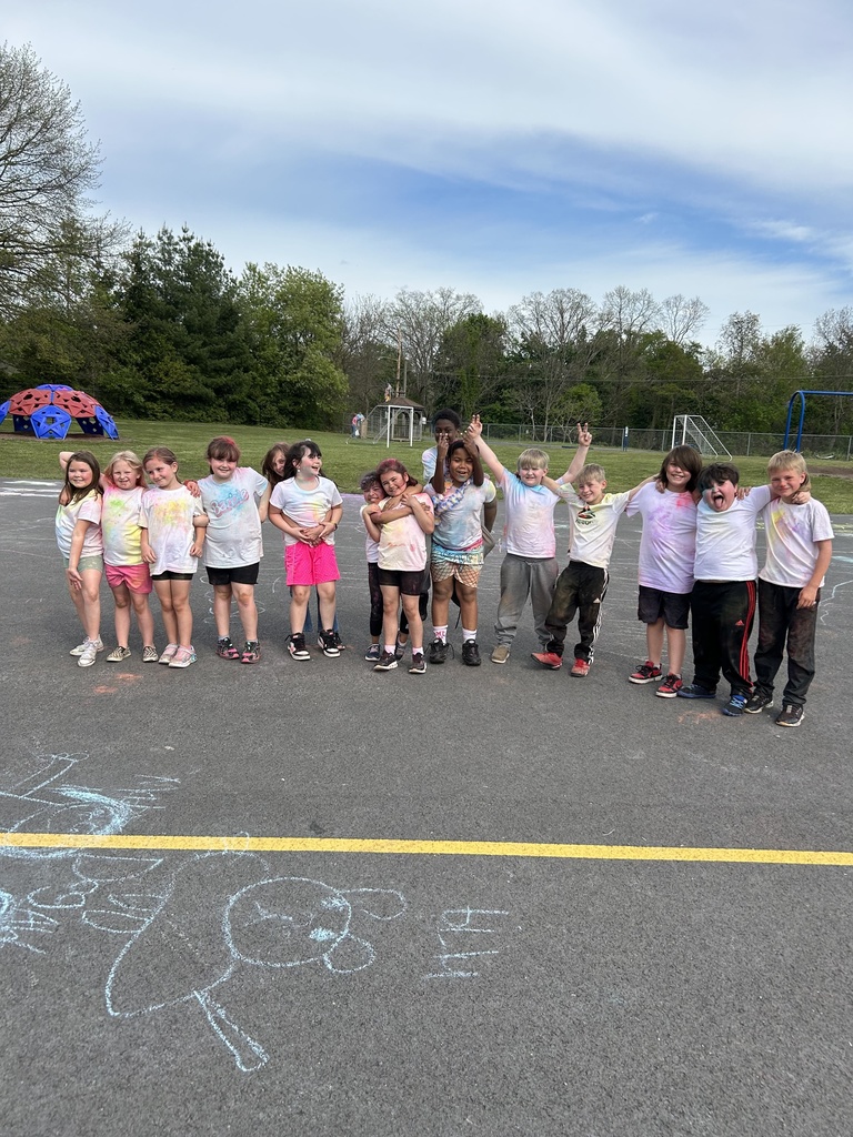 A group of elementary students stand in a line on an outdoor playground blacktop, smiling and posing with colorful powder on their white shirts. Chalk drawings are visible on the pavement in front of them, with trees, playground equipment, and a blue sky in the background.
