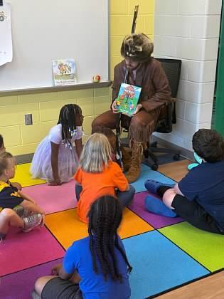 Person dressed in costume reads book to group of children sitting on floor in classroom.