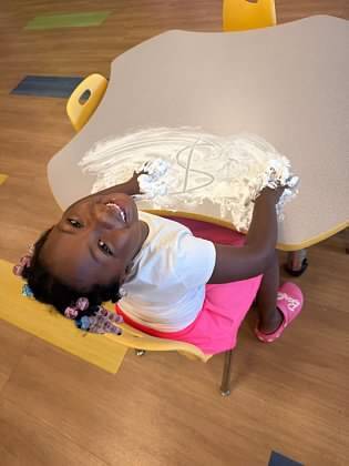 A young child sitting at a table, writing in the air with flour. The table has a white surface.