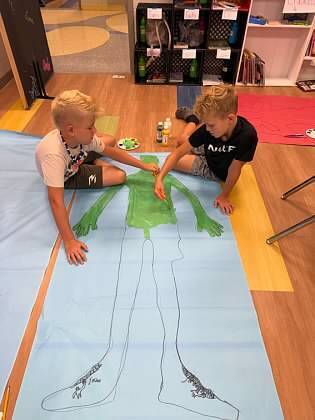 Two children are sitting on a floor, working on a large, green human figure drawn on a blue sheet.