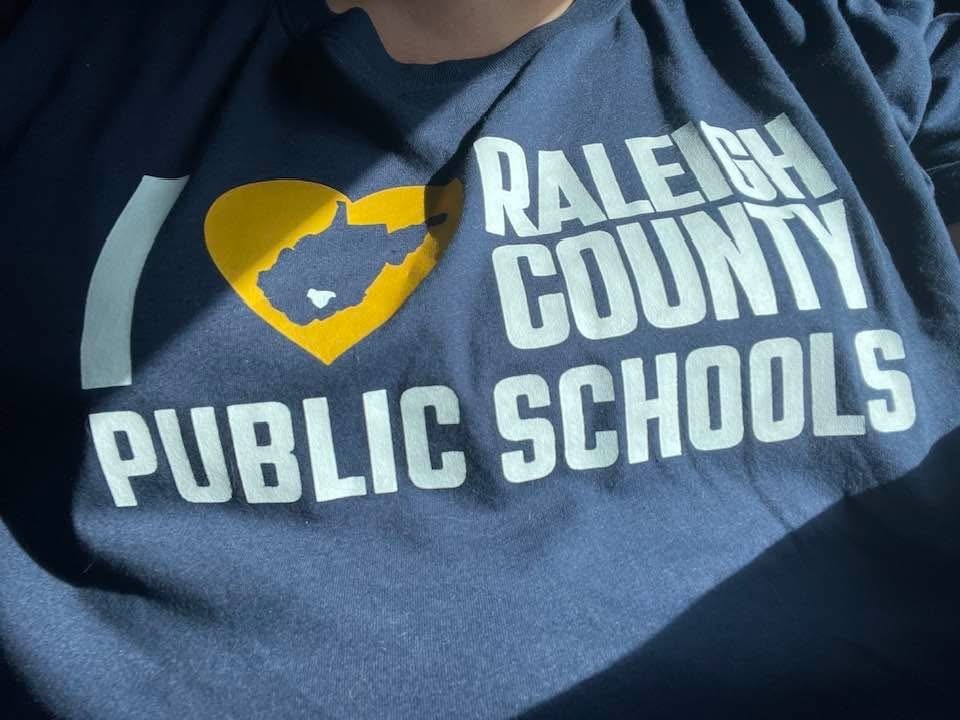 A close-up of a blue shirt featuring "Raleigh County Public Schools" text with a yellow heart shape and state outline.