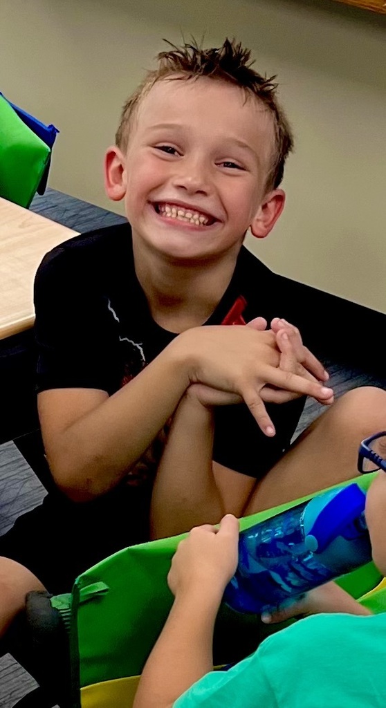 A boy smiles and rests his chin on his hands, sitting near another child. Both are seated on chairs in a classroom.